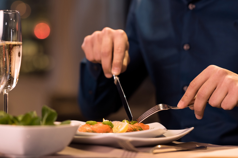 Close up of a male hands cutting and eating delicious salad with knife and fork at a restaurant. A man enjoying a meal at a restaurant. Close up of a plate of salmon fillet at a luxury restaurant