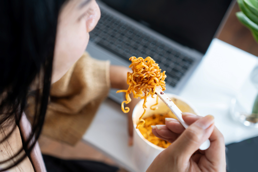 Businesswoman eating spicy noodles at workplace sitting at the office with notebook on desk, unhealthy lifestyle eating fast food concept