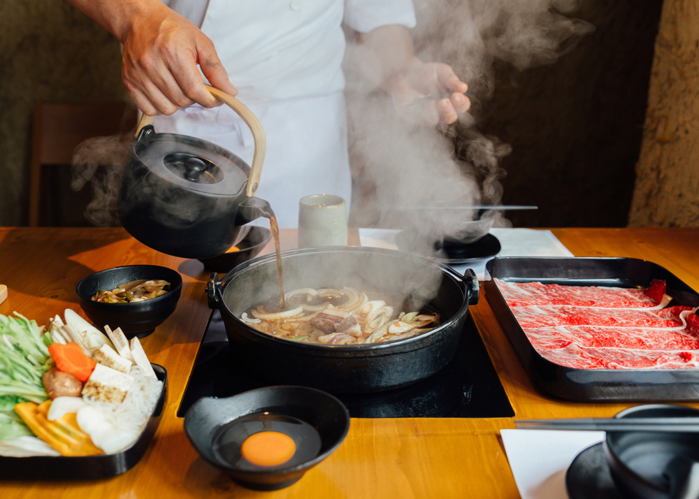 Chef is pouring soy sauce on stir fried vegetables such as onion, cabbage, scallion and tofu and more in hot pot with steam before boiling Wagyu beef