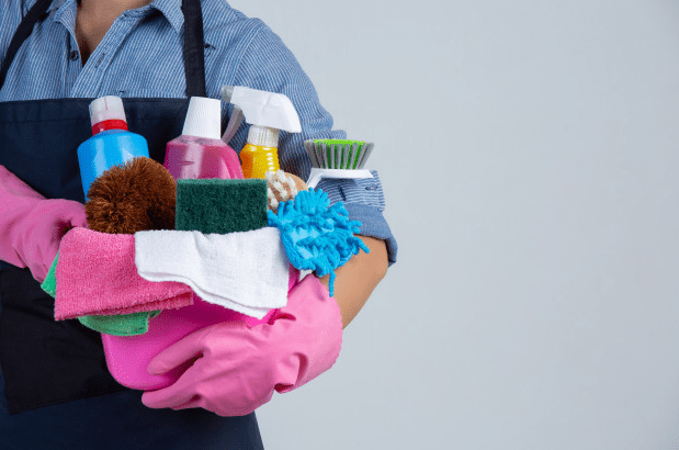 Young girl is holding cleaning product, gloves, and rags in the basin on white wall