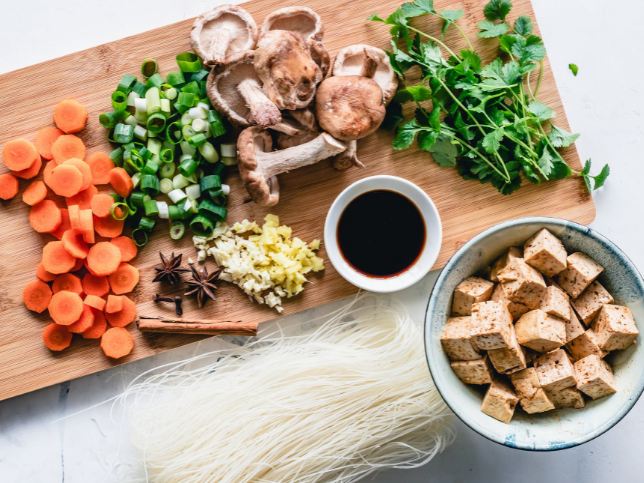 A cup of soya sauce, vegetables, and cubed meat on a cutting board with some noodles