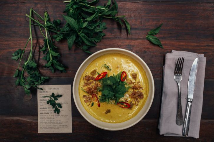 Bowl of soup with some leaves, a receipt, napkin, and a fork and knife on a table