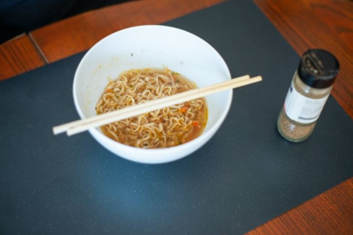 A bowl of soup noodles with chopsticks and a jar full of black pepper
